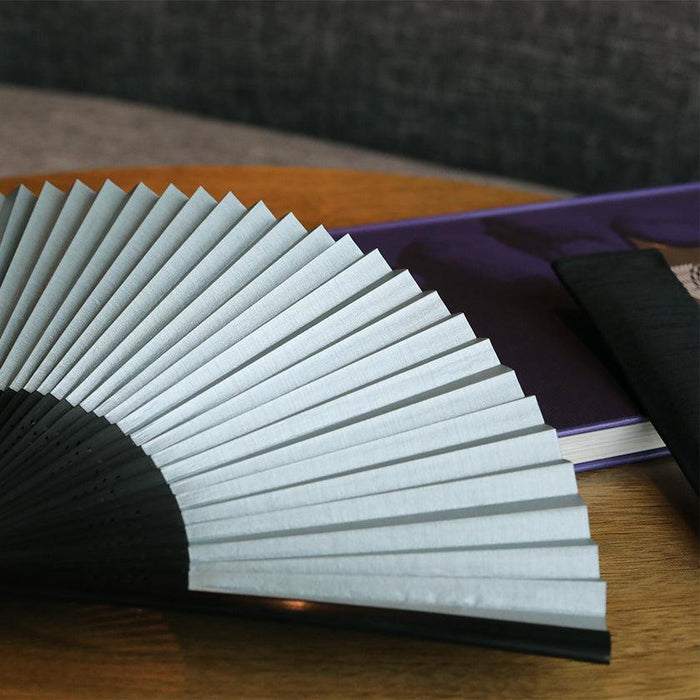Open Japanese folding fan with pale blue pleated paper and black bamboo ribs on wooden surface | Nagoya, Suehirodo